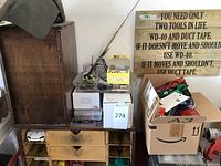 Wooden sign reading 'You need only two tools in life: WD-40 and duct tape: if it doesn’t move and shouldn’t use WD-40, if it moves and shouldn’t use duct tape' leaning against wall; cardboard boxes and wooden drawers with parts