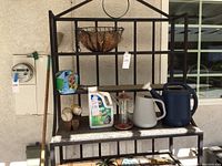 Outdoor black metal shelf with gardening items on three shelves and garden hoe leaning on left.