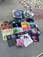 Vinyl records visibly laid out on concrete and brick surface showing various album covers including My Fair Lady, Easy Rider, Hall of Fame Hits and more