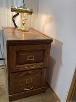 Side angle photo of the wooden filing cabinet showing two drawers with brass handles and label holders, with lamp and power cords visible.