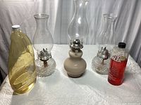 Three vintage glass oil lamps with two bottles of lamp oil on white cloth background