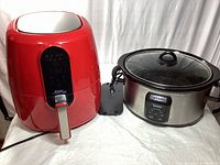 Red Power Air Fryer next to large Crock-Pot slow cooker on white cloth surface, both with electrical cords visible