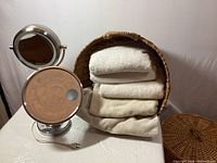 Photo showing one lighted vanity mirror with metal frame and circular reflective glass, large woven basket filled with five white folded towels, and part of another woven basket lid visible.