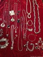 A wide view of assorted silver-tone costume jewelry laid on red velvet including chunky chains, several necklaces with black stone pendants, charm bracelets with multiple charms, and hoop earrings.
