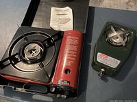 Two portable propane burners on a floor surface, one red Thunder Ranch brand and one green Coleman brand with their stove grates and controls visible, along with a Thunder Ranch instruction manual.