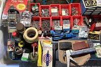 Hardware items on table including multiple red plastic bins filled with various nails, screws, fasteners, loose hand tools, black electrical tape rolls, steel wool and a new poly tarp in plastic packaging.
