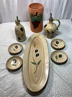 Front view of the olive and wine themed ceramic and terra cotta serving set, showing the oval baguette platter with olive design, two ceramic pitchers with metal pour spouts featuring olive motifs, four matching small dipping bowls, and a terra cotta wine carafe with grapevine decoration.