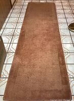 Rust colored wool runner rug shown on tiled floor, solid color with some edge wear visible