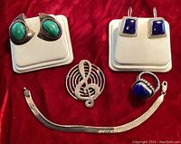 Top view showing the entire set: green malachite earrings, blue lapis lazuli earrings, ring, bracelet, and treble clef pin laid out on red fabric.