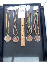 Four beaded necklaces arranged side by side on black tray; ruler in center; two pale yellow citrine bead strings on left with round Buddha medallions, two multi-colored bead strings on right with round Buddha medallions