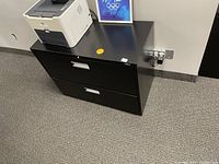 Black 2-drawer filing cabinet seen from angle with printer on top