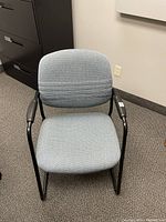 Light blue fabric chair with black metal frame and armrests on carpeted floor in office setting.