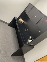 Photo of a black wooden desk with overhead storage against a pale wall and carpeted floor. The desk has two cabinet doors on the overhead storage and the surface shows some wear and tear. Electrical items and miscellaneous objects on the desktop are not included in the lot.