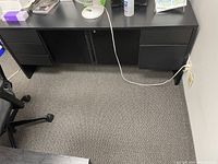 Black hutch with drawers on each side and two central closed cabinets, placed on carpeted floor near walls and outlet with a cord plugged in.