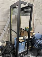Full view of black metal server rack frame with mounting rails and some surrounding items like chairs and bins nearby.