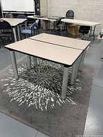 Two beige-top rectangular tables side by side on gray patterned rug with adjustable silver legs visible