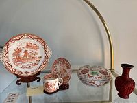Display of three pieces of Asian porcelain pottery on carved wood stands including a platter, cup with saucer, and a lidded ceramic dish, plus carved wax vases