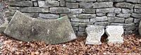 Wide view showing semicircular concrete bench top and two ornate concrete base supports placed on ground with dry leaves, against stone wall.