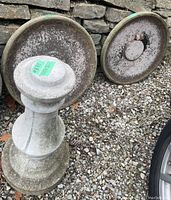 Concrete bird bath base and two tops placed near a stone wall on gravel. Base is approx. 26 inches tall with visible weathering.