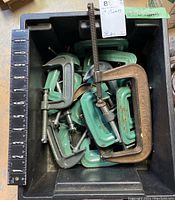 Photo showing multiple C clamps in a black plastic bin with a 12-inch ruler to show scale. Clamps are primarily green with some gray, mixed sizes.