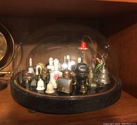 Side view of the glass dome display holding multiple collectible bells arranged on circular black wooden base with wood inlay.