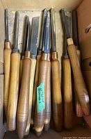 Top view of about a dozen lathe chisels in a box, showing wooden handles and various metal blade types.