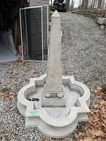 Front view of resin obelisk fountain on quatrefoil base, pump cord laid in basin, set on gravel