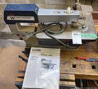 Photo of Ryobi 16-inch scroll saw set on wooden surface including owner's manual and power cord visible.