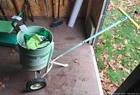 Seed spreader full view showing light green frame, white drum container, wheels, yellow handle grip, in shed setting with tool bench in background.