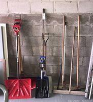 Full lot of shovels and brooms arranged against a cinder block wall, showing red and blue snow shovels, metal shovels, and push brooms