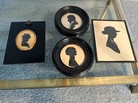 Four framed silhouette art pieces arranged on glass table. Three hand-painted silhouettes in black frames, one silhouette print on board in a rectangular frame.