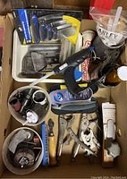 Top-down view showing assorted tools including rotary hole saw bits, hot glue gun, locking pliers, scrapers, spreaders and small hand tools inside cardboard box.