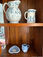 Two shelves showing four ceramic items: two vintage cream and blue pitchers on top shelf; Wedgwood jasperware pitcher and heart-shaped dish in blue and white on bottom shelf.