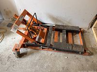 Orange motorcycle jack resting on a concrete floor, showing side view with hydraulic jack and caster wheels.