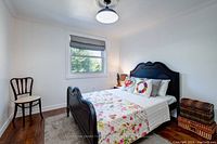 View of antique oak double bed in a room with white walls and brown wood floor. Bed made with floral bedding and two colorful pillows. A wooden chair is placed near the window.