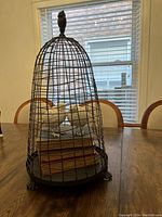 Full view of bird cage with two faux birds perched inside on top of stacked vintage books, displayed on wooden table in front of window.