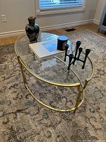 Oval glass top coffee table with decorative items on top and visible gold metal frame and bottom glass shelf.