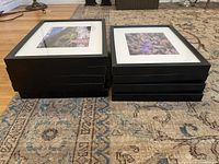 Stack of nine black framed gallery wall prints showing frame thickness and front glass view