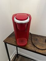 Front view of red Keurig coffee maker placed on wooden table, showing main body, lid, drip tray, and power cord.