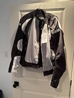 Front angled view of the black, gray, and white motorcycle jacket hanging on a door hook, showing the front zip closure and snap button collar.