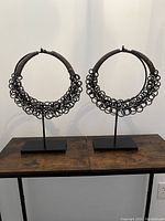 Pair of metal sculptures shaped as large necklaces mounted on black stands, placed on wooden tables against a plain wall.