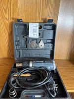 Pro-Fuse heat tool kit open inside black carrying case showing heat tool, attached cord, metal scraper plates, and interchangeable nozzle attachments.