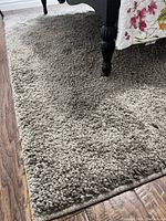 Close-up of light brown plush rug next to wood floor and bed leg, showing thick pile texture and finished edge.