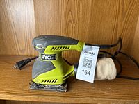 Front and side view of Ryobi corded palm sander with dust collection bag and power cord visible.