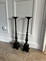 Pair of tall black candle sticks standing on tile floor against white wall, showing full height and bases