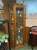Full view of tall oak corner curio cabinet showing glass front door, interior with multiple shelves holding glassware and collectibles, and light inside.