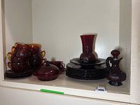 Shelf with stacked cranberry glass plates, candy dish with cover, vase, and Avon Grape Seed Collection cruet