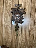 Full view of the wooden cuckoo clock hanging on wood-paneled wall showing the front, carved bird, leaves, weights, and chains.