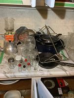 Overview of lot showing cast frying pans, syrup cans, aluminum teapot, glass juicer, salt and pepper shakers and glass containers on countertop.
