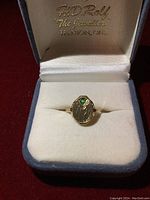 Ring displayed in open jewelry box, top-down view showing oval face with green stone.